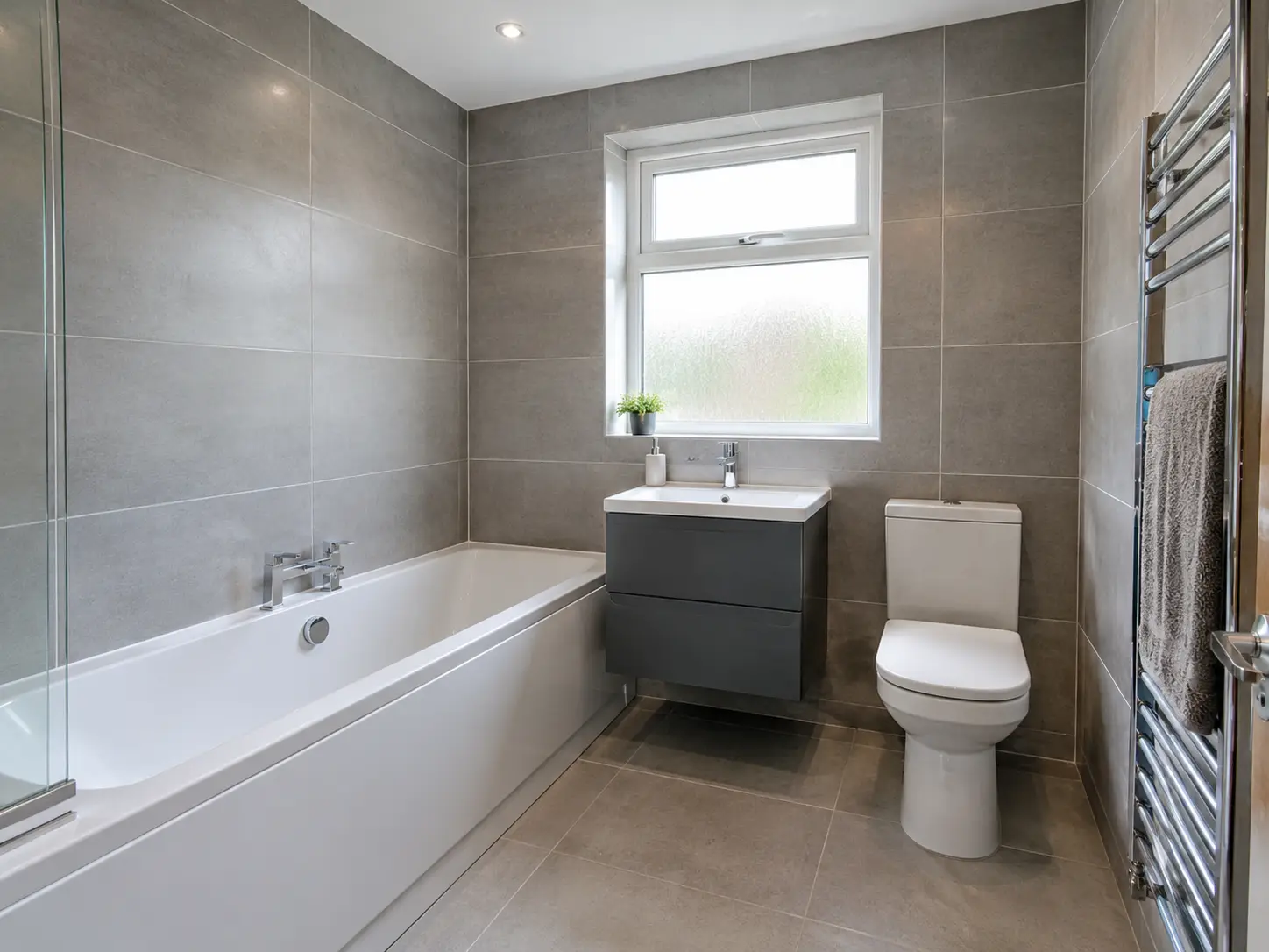 Newly fitted bathroom with large grey wall tiles, white bath, wall-hung vanity and chrome towel rail